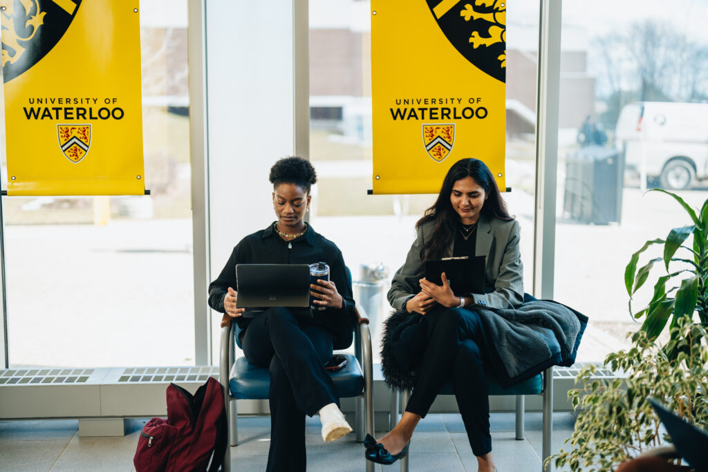 Two students are sitting in front of a University of Waterloo banner