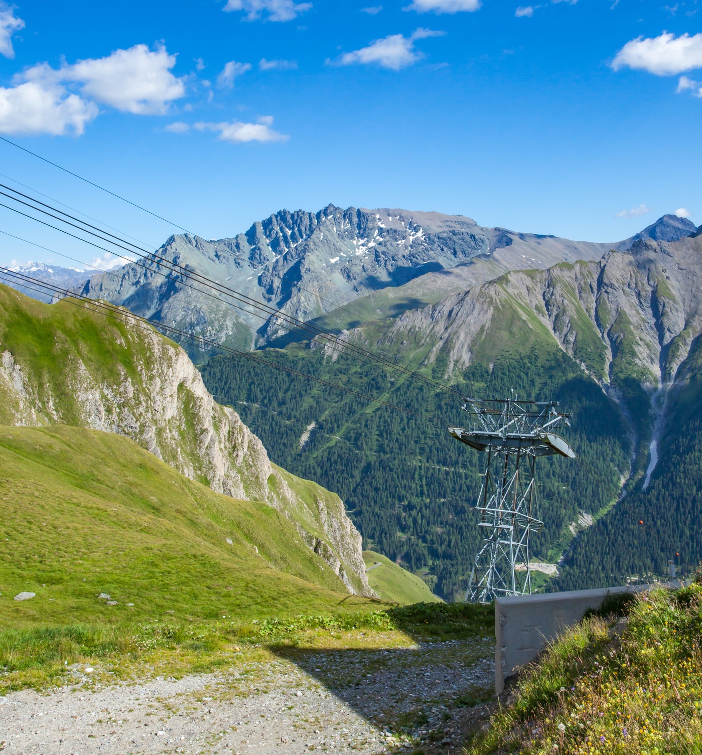 Une tour de téléphérique et des câbles suspendus traversent une vallée alpine spectaculaire, avec des montagnes enneigées, des prairies verdoyantes et des pentes boisées sous un ciel bleu parsemé de nuages blancs.