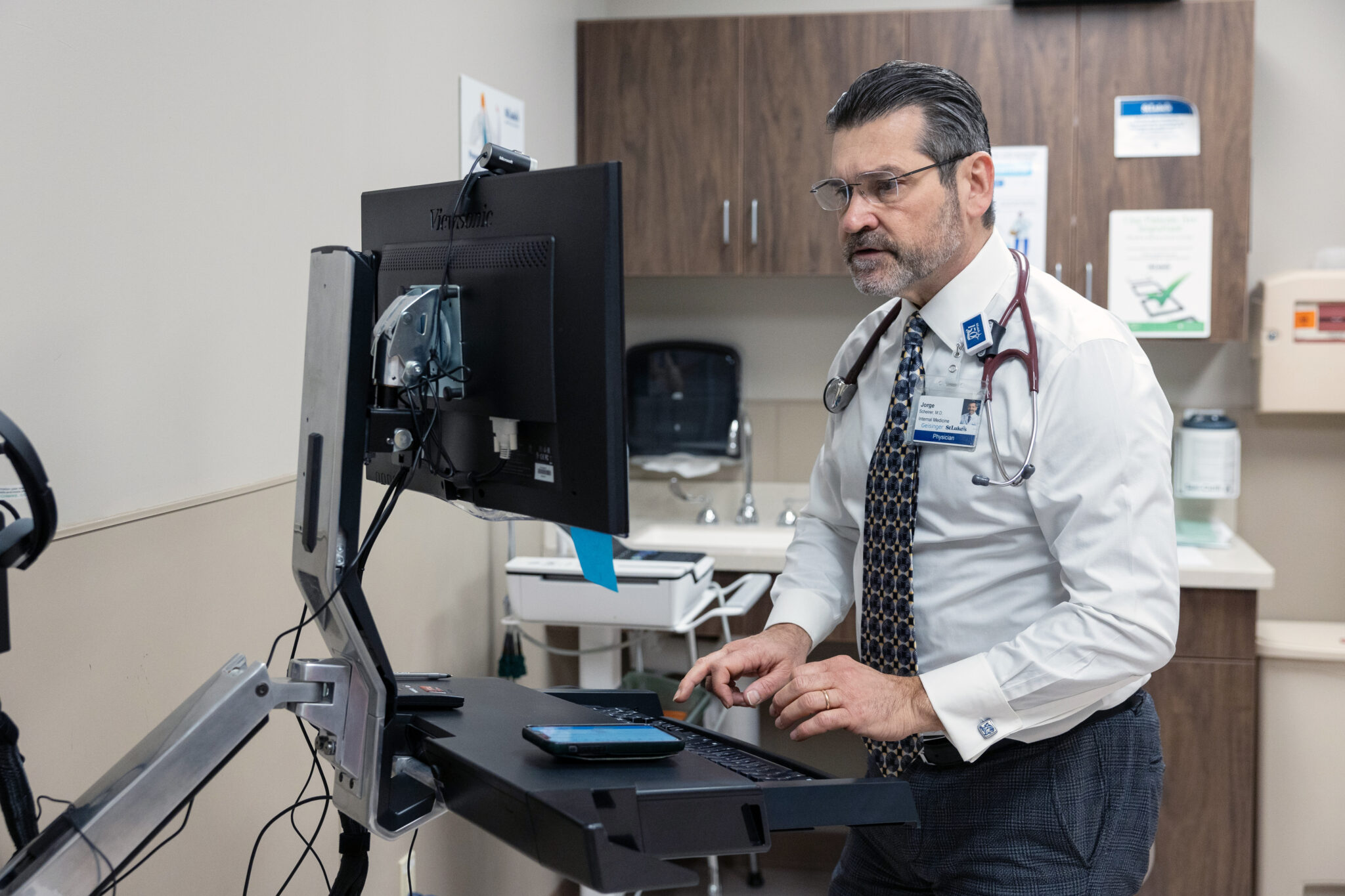 Un médecin en sarrau blanc avec un stéthoscope est assis à un bureau debout, utilisant un moniteur d’ordinateur dans un cabinet médical.