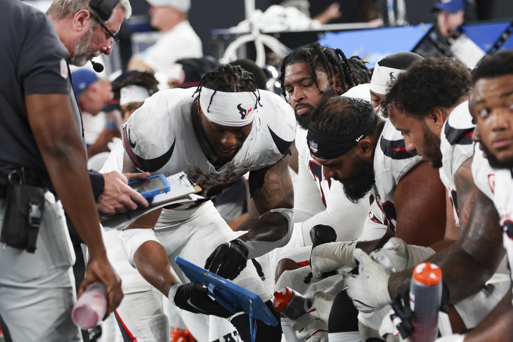 The image shows a group of football players wearing white jerseys with black and red accents sitting on a bench. They are engaged in an activity, with one player holding a blue tablet device and others appearing to be focused on it. A person wearing a dark shirt and pants is standing nearby, holding a clipboard and a pink water bottle. The background includes blurred elements, such as equipment and other individuals, with a blue triangular structure visible. The players are wearing gloves, and some have headbands or braided hairstyles.