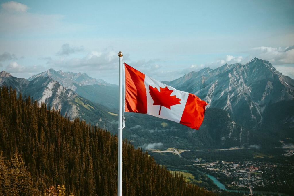 Drapeau canadien flottant sur un mât devant un paysage montagneux, des forêts denses et une petite ville dans la vallée sous un ciel partiellement nuageux.