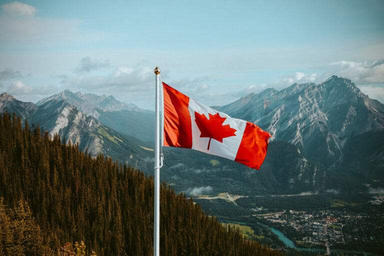 Canadian flag flying on a flagpole with a mountainous landscape, dense forests, and a small town in the valley under a partly cloudy sky.
