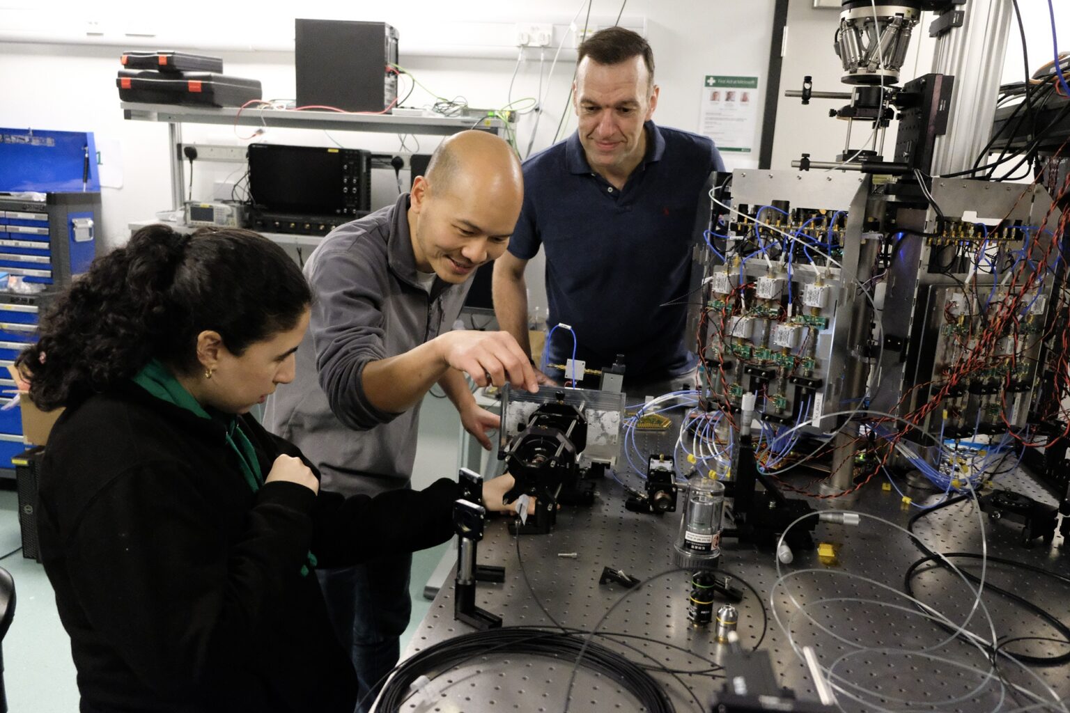 A woman, at left, and two men standing next to a large table covered with complex machinery and cables and wires. They are looking at it closely